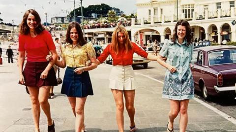 Alternative description
Four women link arms on a seaside promenade and smile at the camera. In the background are steps leading down to a beach.