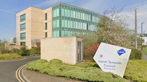 North Tyneside Council's four-storey building. It is mostly made of glass. There is grass and trees in front of it and a grey sign reading North Tyneside Council.