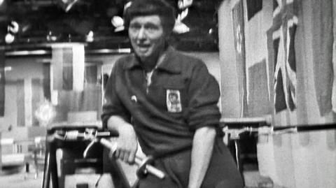 Black and white image of Blue Peter presenter John Noakes holding a small dumbbell and looking out of breath.