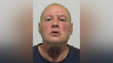 Mugshot of Straughan. He is bald with a cut above his left eye and a large red rose tattooed across the front of his neck