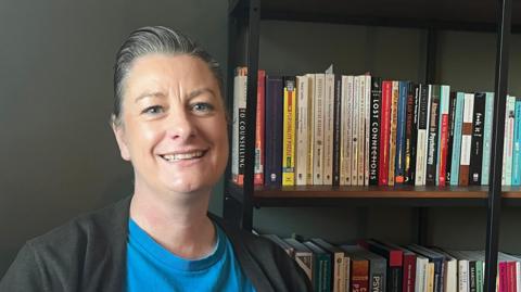 Zoe Hughes, an Exeter City council member, wearing a blue T-shirt and a black top, sitting next to a bookshelf