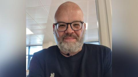 A selfie of a bearded and bespectacled man who smiles at the camera. He is wearing a blue top with a white logo on his chest.