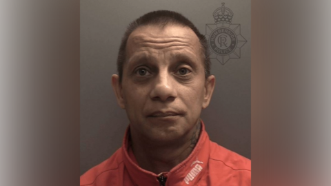 A police custody photo of Patrik Seidenglanz, who has short brown hair and a tattoo on the side of his neck. He is wearing a red Puma zip-up jacket. The wall behind him is grey. 