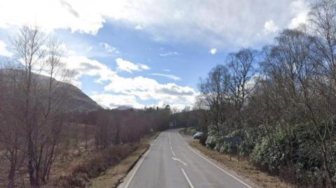 A road stretching ahead with trees of either side. Mountains can be seen in the distance.