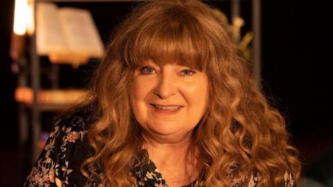 Janey Godley looks into the camera smiling. She has long curly brown hair with a fringe and is wearing a black floral blouse