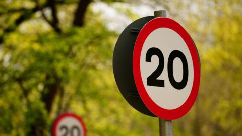 A red 20mph speed sign. Green trees are behind the sign.