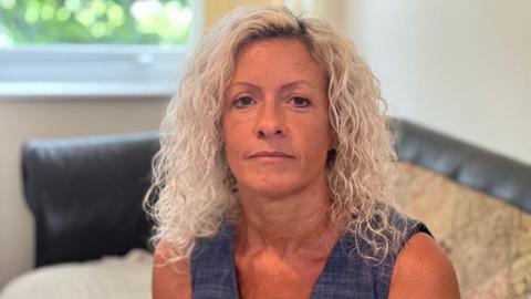 Michelle sat on a sofa looking at the camera. She has shoulder-length blonde curly hair, blue eyes and has a stud nose piercing. 
