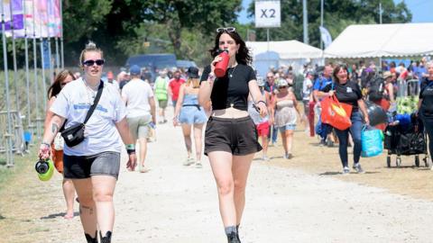Festivalgoers arrive at the Isle of Wight Festival at Seaclose Park on June 19, 2025 in Newport, Isle of Wight two women can be seen walking along a dusty path with crowds behind them. one is drinking a bottle of water