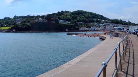 The seafront of Teignmouth, a small south Devon town. There is a promenade, with blue railings, a small beach, and a green headland with trees and buildings on it.