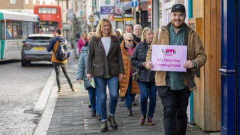 A group of people walking in a tour group, following a man with a cap on and holding up a sign saying "Love Grimbsy: NEL FOOD TOUR MEETING POINT"