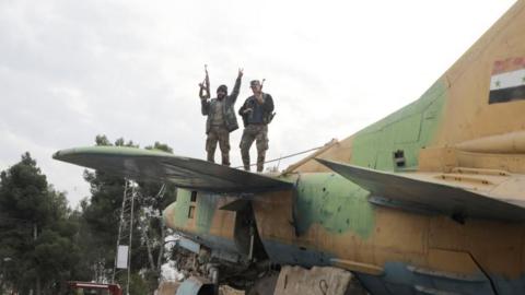 A rebel fighter gestures the victory sign while standing on a military aircraft that belonged to forces loyal to Bashar al-Assad government, inside Hama's military airport, December 7, 2024