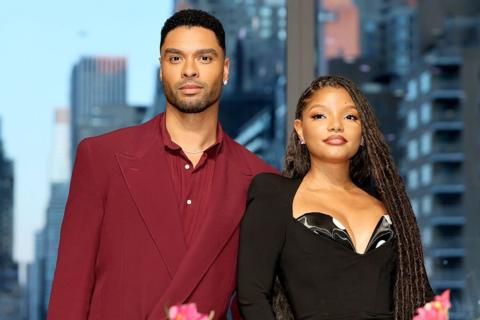 A man in a burgundy suit with black hair is looking into the camera. Next to him is a woman in a black suit dress and long braided hair. There is also a background of blue skies and multiple skyscrapers.