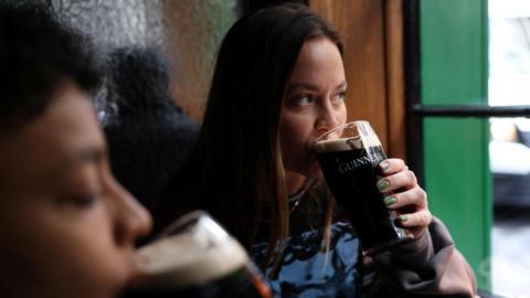 A woman with dark hair drinks a pint of Guinness beer in a pub while looking out of green-lined window.