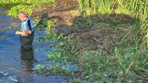 Steven Hillier, in waders, standing in water by a river bank with a metal object with fins, roughly the size of a wine bottle, sitting on trodden down vegetation behind him
