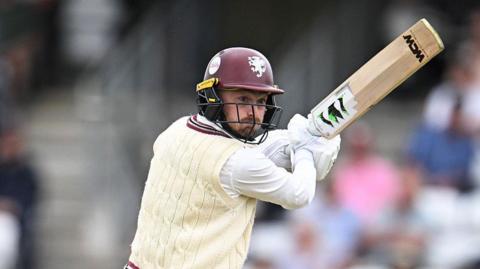 Lewis Goldsworthy in white cable jumper and maroon batting helmet holds his bat up to his right as he plays a shot