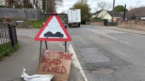 Road with a pothole and a sign calling for the road to be fixed