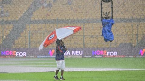 A man stands on a cricket field, holding an umbrella under heavy rain and looking up at a spider camera device
