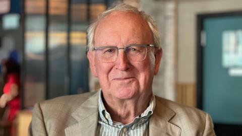 Tony Beckingsale is pictured wearing a cream jacket and a green and white striped shirt. Mr Beckingsale has white hair, is wearing glasses and is smiling. The background is blurred.