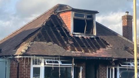 A shot of a traditional brick house with a terraced roof which has been significantly damaged by fire. Many of the roof's tiles are completely missing and a box window in the attic has been blackened and warped. 