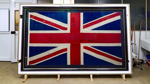A large framed union jack flag is displayed upright indoors, leaning on wooden supports in what seems to be a workshop or conservation space. The flag shows visible creases, small marks, and wear. The red, white, and blue design fills most of the frame while tools, and equipment are visible in the background.