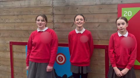 Three schoolgirls wearing red jumpers and grey skirts standing in front on a wooden panelled wall. On the wall, there are boards with numbers on them in colourful shapes.