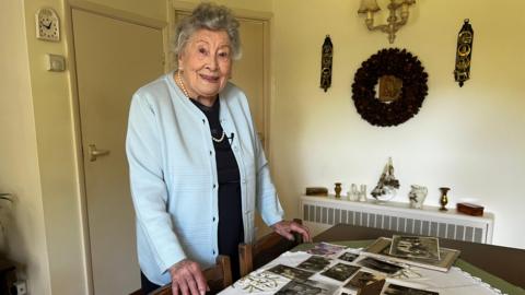 Silvia Verrall looks at the camera and smiles, she is standing next to a table covered in old photographs and is wearing a pale blue jacket