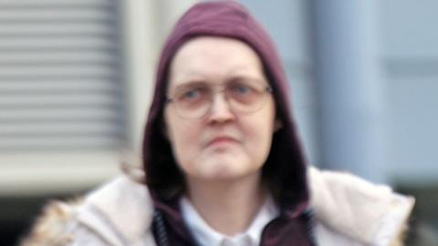 A transgender woman, with a stern expression wearing glasses and a burgundy hoodie which is covering her head.