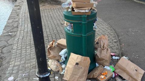 A green bin surrounded by litter, specifically brown paper bags from a local fish and chip shop and other takeaway boxes.