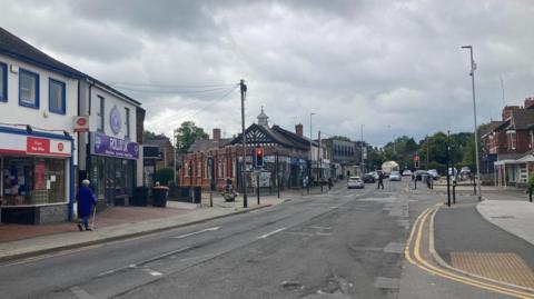 A busy town street with shop buildings of varying ages on either side, traffic in both directions and pedestrians walking down the pavements.