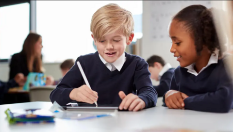 Two children sit at a table in a classroom with a boy writing something and a girl looking down at what he is doing. The boy has short blonde hair and the girl brown hair tied back. Both are wearing a blue uniform with a white shirt. People can be seen blurred in the background.