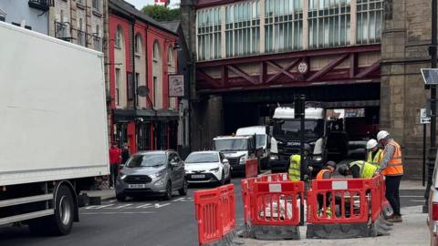 A busy road with a railway bridge overhead and men in orange jackets and white protective helmets by the roadside