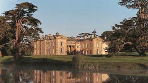 A large stately looking building that appears to be made of sandstone is surrounded by trees, with a body of water in the foreground