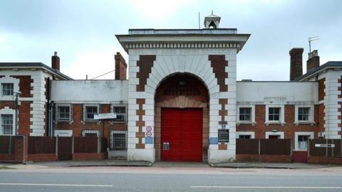 The exterior of HMP Aylesbury. There is a large red door and the rest of the building is made of brick and white stone. 