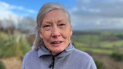 An older woman with shoulder-length grey hair, wearing a grey zip-up fleece. She is looking at the camera with a neutral expression. The background behind her is out of focus, but she appears to be stood outside with green fields behind her.