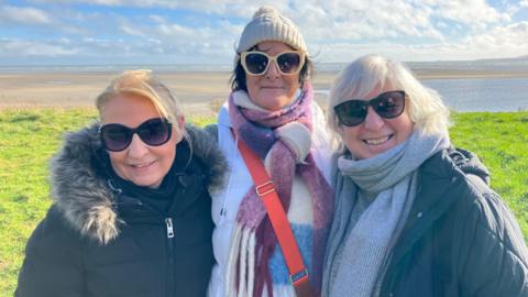 Three women wear sunglasses. They stand with grass and a beach behind them. The sky is blue with some white clouds. They are wearing coats, and scarves. The woman in the centre is wearing a white wooly hat.