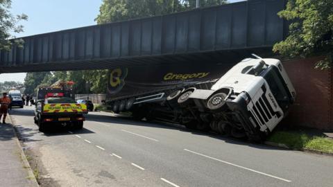 A lorry wedged under a blue railway bridge, with its trailer partially crushed. Highways officers are parked nearby.