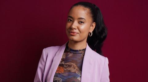 A woman stands against a red backdrop. She is wearing a pink suit jacket and a multi-coloured top. She has straight black hair.