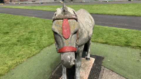 A concrete sculpture of a donkey with red harness on its face. The statue stands on a plinth on a green lawn