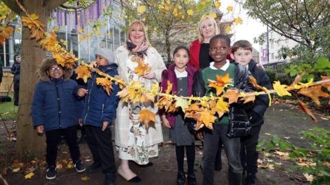 Mayor of Sunderland Allison Chisnall cuts the ribbon to open Mowbray Park Community Garden. She is standing with councillor Beth jones and five school children. The ribbon has been decorated with autumnal leaves.