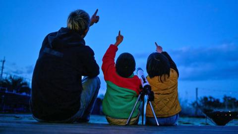 Silhouette of father and two children pointing to the night sky. They have a telescope beside them.