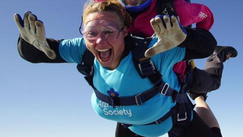 Kaye mid-air skydiving wearing a blue Alzheimer's Society shirt and has white and blue gloves on. She's also wearing goggles. She's strapped to another person who is above her and leading the skydive. 