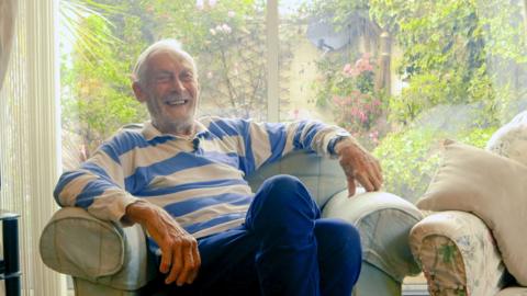 A smiling Nigel sitting on an armchair. He is wearing a blue and white checked rugby shirt and blue trousers. Behind him is a garden with roses. 