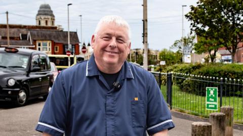 Tony Norman, who wears his nurse's blue uniform top, smiles as he poses on a pavement. A black cab and an old building is in the background.
