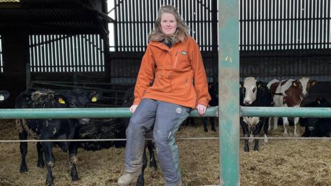 Caitlin Riddell is sitting on a bar in front of some young cattle. She's a young woman with shoulder length fair hair and wearing an orange top and waterproof trousers 