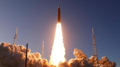 The Space Launch System rocket lifts off from the Kennedy Space Center in Cape Canaveral, Florida. There are brilliant white flames and lots of smoke around pylon-type structures.