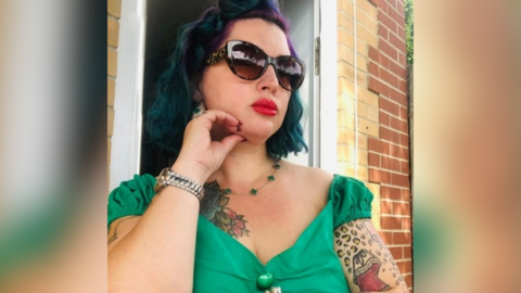 A head and chest image of Tania Williams. She is wearing green short-sleeved dress and is resting is touching her chin with her right hand. She has dark, dyed hair, sunglasses and tattoos on her upper arms and on the right of her chest. She appears to be posing outside the door of a house.