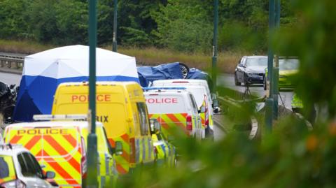 An overturned car is seen under a tarp, surrounded by police vehicles on a road.