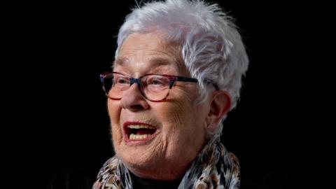 Kathy Martin, 80, portrait, in front of a black background, laughing at something off camera