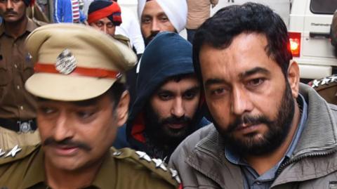 Jagtar Singh Johal being escorted to a court in Ludhiana on 24 November 2017. He is wearing a dark blue hooded top with the hood up. Two men are in the foreground, one in a uniform and ther othey in a grey jacket, and there are a number of people in the background.