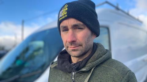 A man who is wearing a black beanie hat is looking at the camera. He's wearing a green fleece with a black snood around his neck. He is standing in front of a white van.